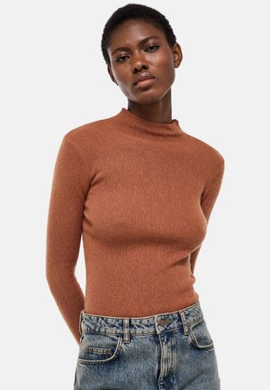 Femme portant un pull à col roulé côtelé ajusté de couleur rouille et un jean taille haute bleu clair, debout devant un fond blanc uni.