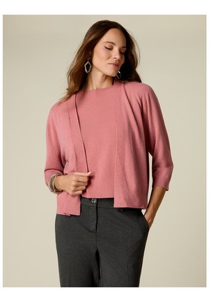 Vrouw die een roze cardigan en top draagt met donkergrijze broek, kijkt naar beneden met één hand in de zak en de andere hand houdt de cardigan vast.