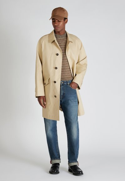 Hombre vestido con un abrigo beige, suéter marrón con patrón, jeans azules con puños, zapatos negros y gorra marrón, de pie contra un fondo gris.