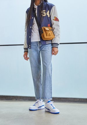Osoba v modrobílé univerzitní bundě, světle modrých džínách, bílých a modrých teniskách a s malou hnědou kabelkou přes rameno.