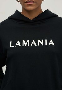 Juodas miesto stilius su priekyje esančia kišenė ir dideliu baltonu tekstu "LAMANIA" krūtinės srityje, pagamintas iš minkštos, šiurkščios medžiagos.