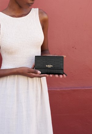 Donna in abito bianco senza maniche che tiene in mano un portafoglio in pelle nera testurizzata con logo dorato davanti a una parete rossa attenuata.