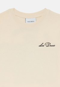 Cremfarbendes T-Shirt mit kurzen Ärmeln aus weicher Baumwolle, mit einem runden Kragen und dem in schwarzer, geschwungener Schrift auf der Brust gestickten "Les Deux"-Logo.