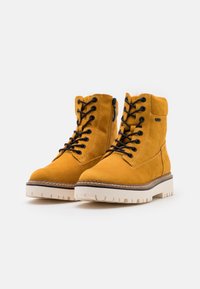 Stivali alla caviglia in suede giallo con lacci neri, zip laterale e suola spessa bianca. I dettagli delle cuciture aggiungono texture alla parte superiore.