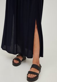 Marineblå plisseret maxikjole med en sideslids, vist med sorte slide-sandaler. Stoffet ser let og tekstureret ud.