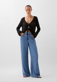 Svart, löst stickad långärmad topp med djup V-ringning och knytdetalj fram, ihop med vida ljusblå denimjeans.