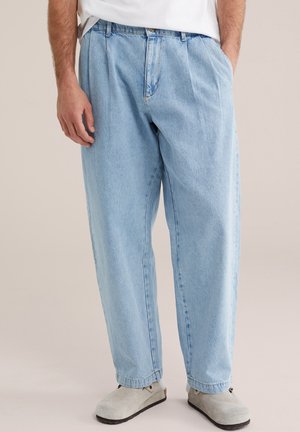 Man draagt losse lichtblauwe denim jeans, wit overhemd en lichtgrijze slip-on schoenen, staat met handen in de zakken.