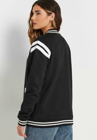 Kvinde med langt bølget hår iført en sort varsityjakke med hvide striber på skuldrene og ærmerne, parret med blå jeans, vendende sig sidelengs.