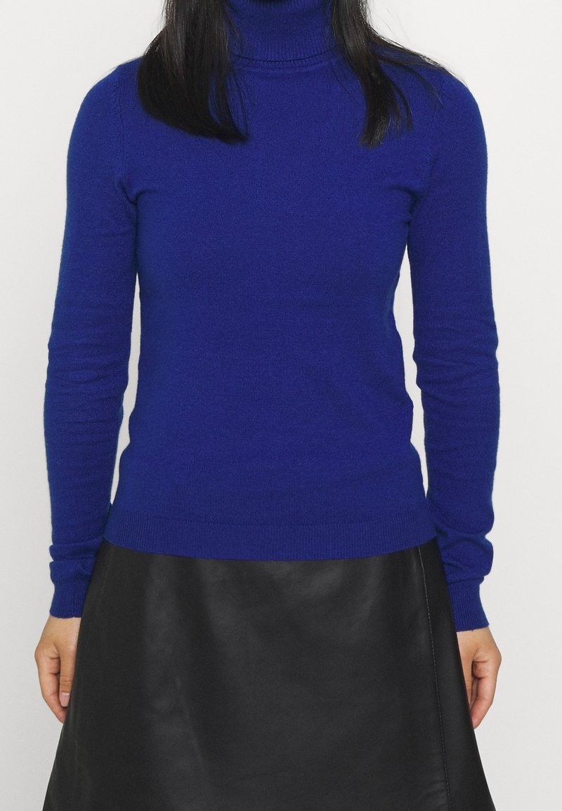 Blå polotröja i mjukt tyg med långa ärmar, kroppsnära passform och ribbade ärmslut och fåll, kombinerad med en svart läderkjol.