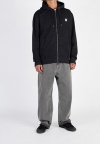 Black zip-up hooded sweatshirt with a front pocket and gray drawstrings, paired with loose gray jeans and black sneakers on a plain background.