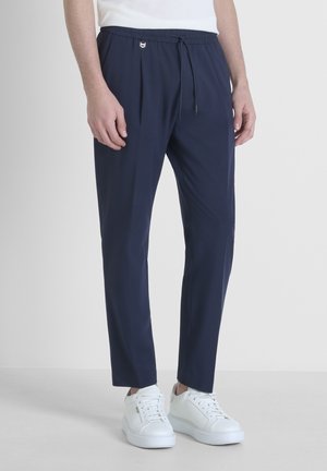 Mann trägt dunkelblaue Hose mit Kordelzug und weiße Turnschuhe, steht vor schlichtem hellem Hintergrund.