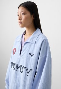 A woman with long black hair gazes to the side, wearing a light blue Fenty jacket decorated with logos and a purple inner layer.