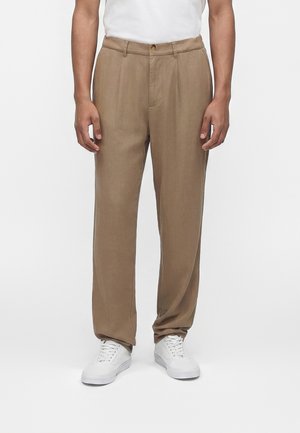 Person iført beige plisserte bukser med beltehemper og hvite sneakers, stående mot en ensfarget hvit bakgrunn.