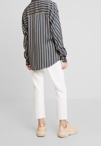 Gestreiftes Hemd in Marineblau und Gelb, aus weichem Stoff, kombiniert mit weißen Hosen. Das Outfit wird von lässigen Sneakern in Beige ergänzt.