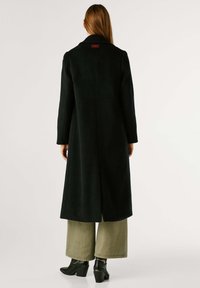 Lungo cappotto nero realizzato in tessuto texturizzato, con colletto a falde e spacchi sul retro. Indossato sopra pantaloni larghi verde oliva e stivaletti neri con tacco.