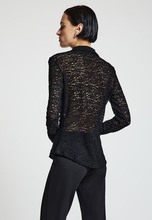 Chemise en dentelle noire avec un motif texturé, manches longues et ourlet légèrement arrondi. Portée sur un pantalon noir, dévoilant des détails translucides.