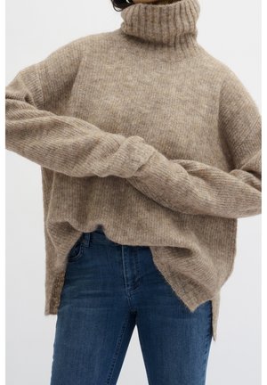 Personne portant un pull à col roulé côtelé beige ample et un jean bleu, les mains croisées à la taille.