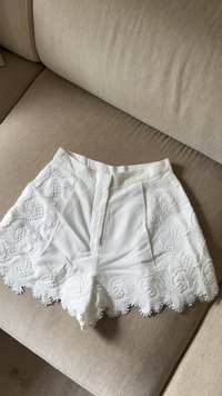 Pantaloni bianchi realizzati in tessuto con intricate ricami a scallop e motivi in rilievo lungo l'orlo, caratterizzati da una piega frontale e una vita.