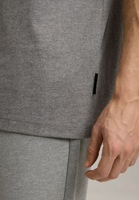 Szary bawełnowy t-shirt o prostym kroju, z czarną metką na bocznej szwie. Ręka trzymająca dół, ukazująca szczegóły tekstury.