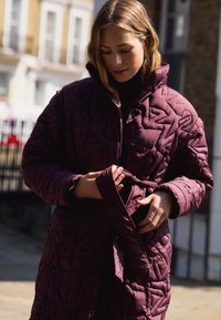 Piumino trapuntato color bordeaux con colletto alto e motivo geometrico testurizzato. Presenta una chiusura anteriore con bottoni a pressione e un dettaglio in vita con lacci.