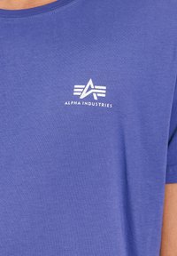 Kék pamut póló kerek nyakkal és rövid ujjakkal. Fehér "Alpha Industries" logó emblémával a bal mellrészen. Sima textúra.