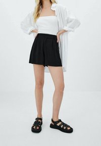 Pantaloni corti neri a vita alta con pieghe, abbinati a un top aderente bianco a coste e a una camicia bianca oversize con bottoni. Sandali neri con cinturini.