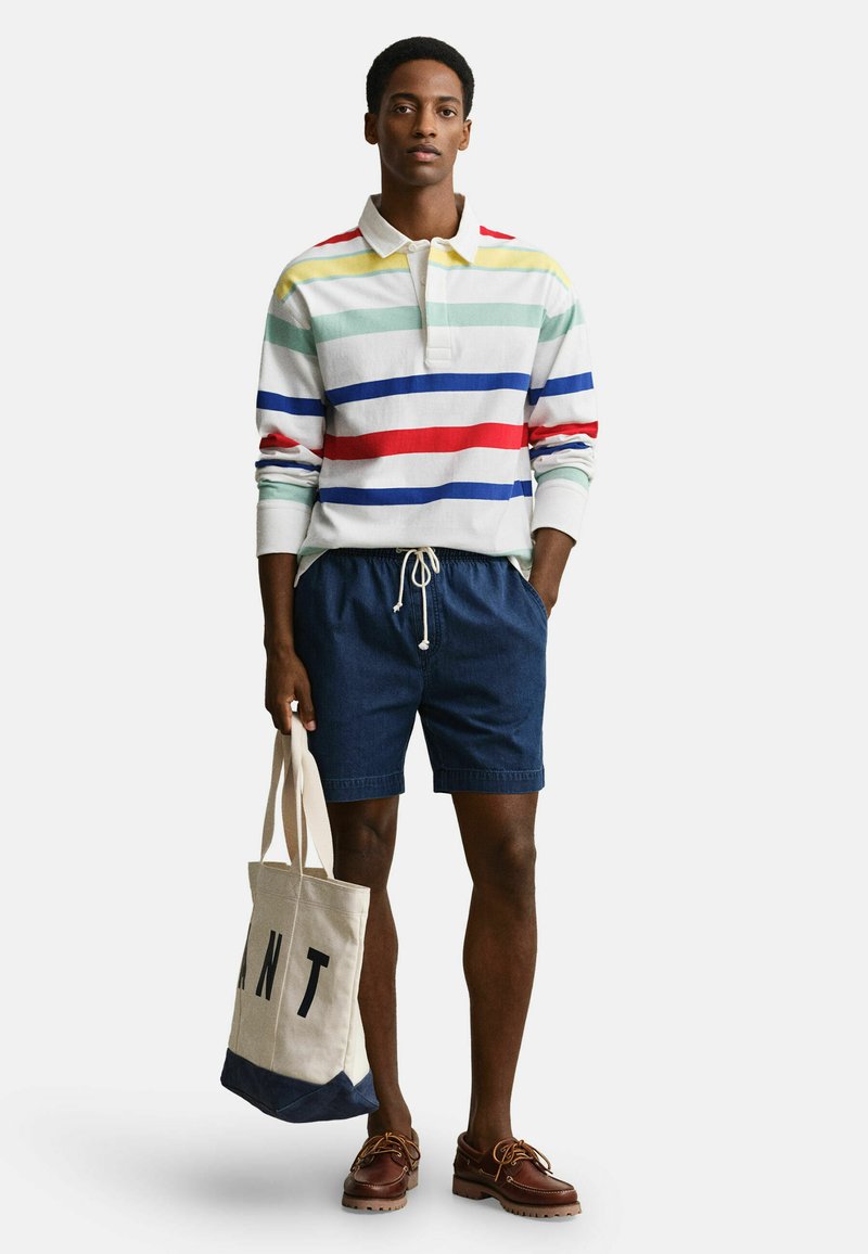 Uomo che indossa una maglia a maniche lunghe a righe multicolori, pantaloncini navy con coulisse, scarpe da barca marroni, che tiene una borsa tote beige con scritte nere.