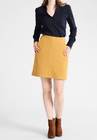 Chemise bleu marine à boutons, associée à une jupe jaune moutarde de longueur genou, avec un tissu texturé et deux poches latérales. Terminée par des bottines brunes à la cheville.