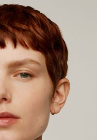 Pendiente de aro dorado con acentos verdes, mostrado en una modelo con cabello corto, texturizado y castaño rojizo, y piel clara, presentando un primer plano de su rostro.