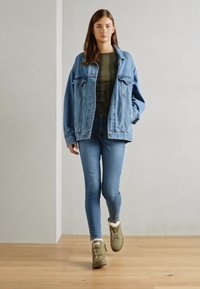 Denimjakke med to frontlommer, grønn camouflasje topp, lyseblå skinny jeans og olivengrønne snøresko med pelsfor.