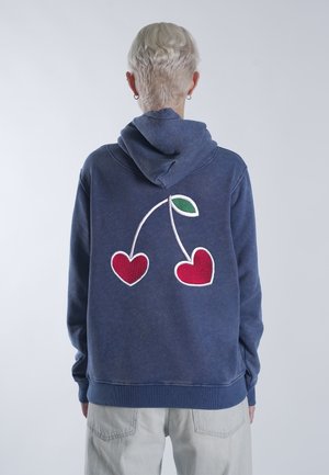 Persoon met kort blond haar die een blauwe hoodie draagt met op de achterkant twee rode kersen in de vorm van een hart met een groen blad.