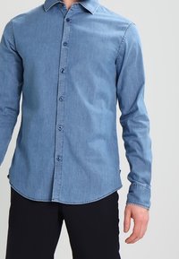 Ljusblå denimskjorta med spetsig krage, långa ärmar, knappstängning fram och rundad nederkant. Knappar i en kontrasterande mörk färg.