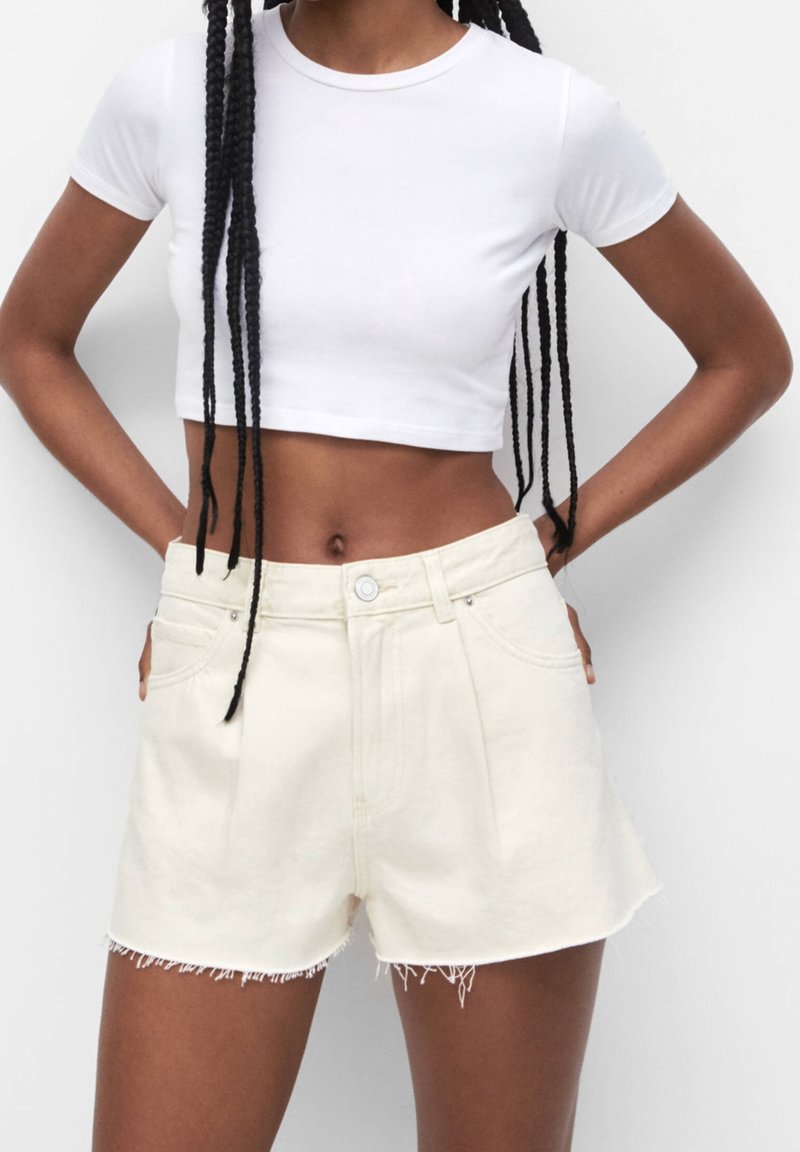 Femme portant un t-shirt blanc court et un short en denim écru taille haute avec des bords effilochés, debout les mains dans les poches.