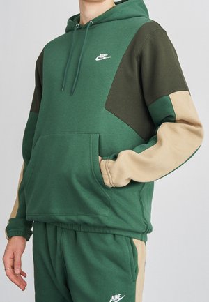 Person trägt einen grünen, dunkelgrünen und beigen Nike-Hoodie und passende Jogginghose mit einer aufgesetzten Tasche vorne.