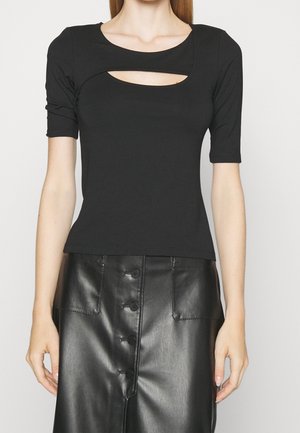 Femme portant un haut noir à manches courtes avec une découpe sur la poitrine et une jupe en cuir noire taille haute à boutons.