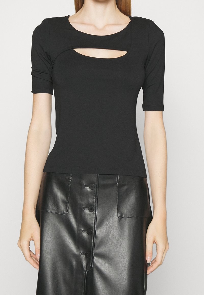 Žena v černém krátkém tričku s výstřihem na hrudi a černé kožené sukni s vysokým pasem a knoflíky.