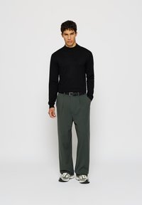 Zwarte gebreide trui met een hoge hals, gecombineerd met loszittende groene broek. Schoenen hebben een grijs en zwart ontwerp. Eenvoudige, moderne stijl.