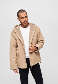 Beige hettejakke med et glatt ytterskikt, to sidelommer og indre fleecefôr, med glidelås foran og justerbar hette.