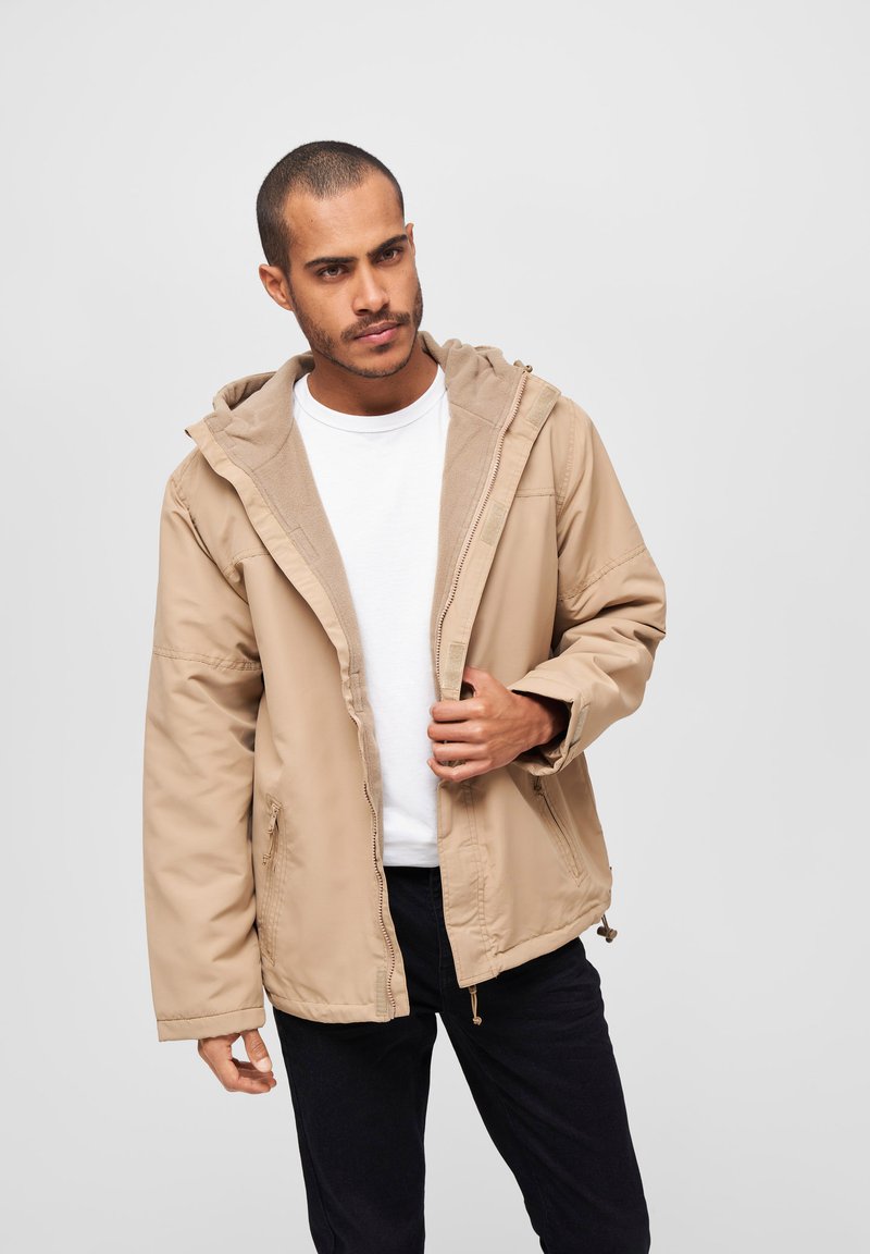 Beige hettejakke med et glatt ytterskikt, to sidelommer og indre fleecefôr, med glidelås foran og justerbar hette.