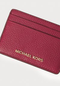 Rød teksturert skinnkortholder med gullprent "MICHAEL KORS"-logo, med flere spor for kort og et elegant design.
