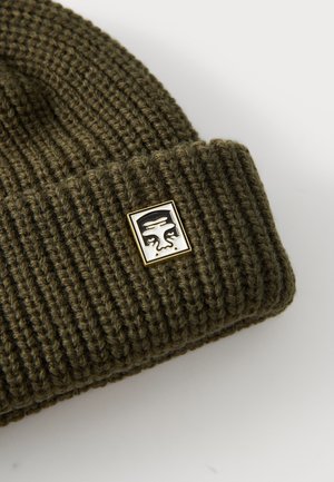 Olive green knit beanie with ribbed texture; features a small square gold and black logo patch on the front.
