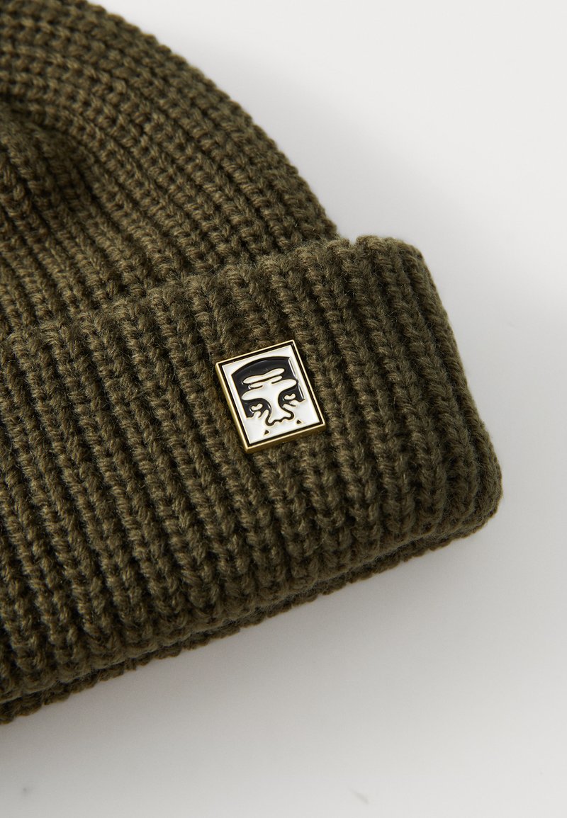 Olive green knit beanie with ribbed texture; features a small square gold and black logo patch on the front.