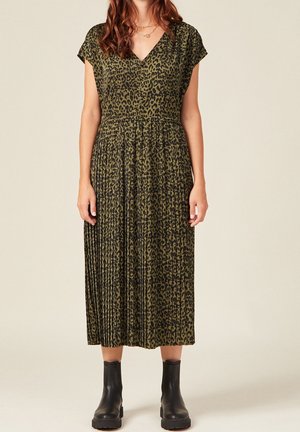 Kobieta ubrana w plisowaną sukienkę midi w oliwkowym kolorze z nadrukiem w cętki, z krótkimi rękawami i czarnymi botkami, stojąca na jasnym tle.