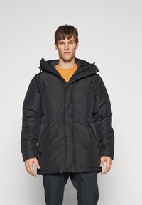 Parka nera imbottita con cappuccio, dotata di zip frontale, coulisse all'orlo e tasche laterali. Il materiale sembra liscio e spesso.