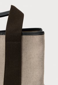 Texturovaná béžová taška s tmavě hnědým popruhem a černým lemem. Disponuje strukturovaným tvarem a viditelným šitím podél okrajů.