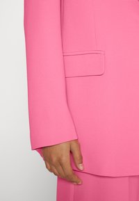 Blazer rosa ajustado com uma textura suave, apresentando um único bolso e mangas ligeiramente afiadas. Vista em close com uma mão pousada sobre ele.