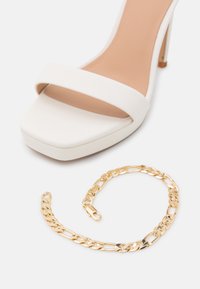 Sandale blanche à talon haut avec bout carré et une seule bride, associée à une chaîne de cheville en or sur fond blanc.