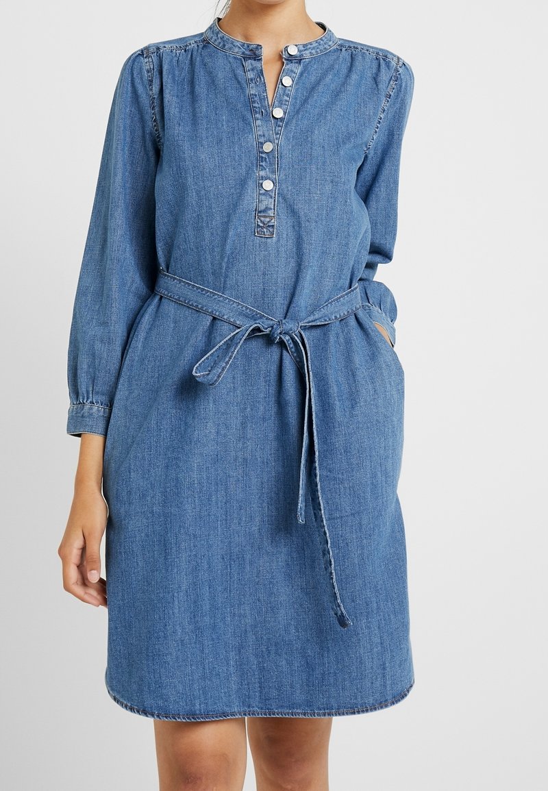 Femme portant une robe en denim bleu à manches longues, col boutonné, ceinture nouée à la taille et mains dans les poches latérales.