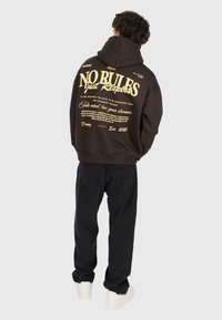 Homme portant un sweat à capuche noir avec un grand texte beige "AUCUNE RÈGLE Juste du Respect" et d'autres phrases motivantes imprimées au dos, pantalon noir, chaussures blanches.