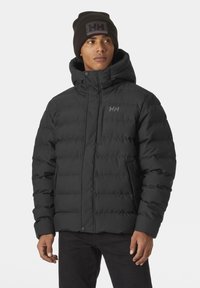 Chaqueta de plumas negra con cremallera frontal, textura acolchada, capucha y logotipo bordado. Combinada con un gorro de punto negro a juego.